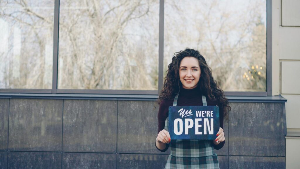A smiling small business owner next to a sign promoting their participation in a community solar program. — community so…